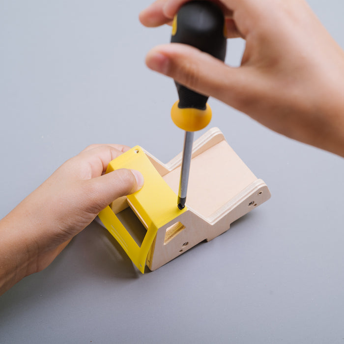 A person uses a screwdriver to assemble the Stanley Jr. Dump Truck Kit by Red Toolbox, featuring a yellow plastic part on a gray surfaceโperfect for kidsโ crafting fun.