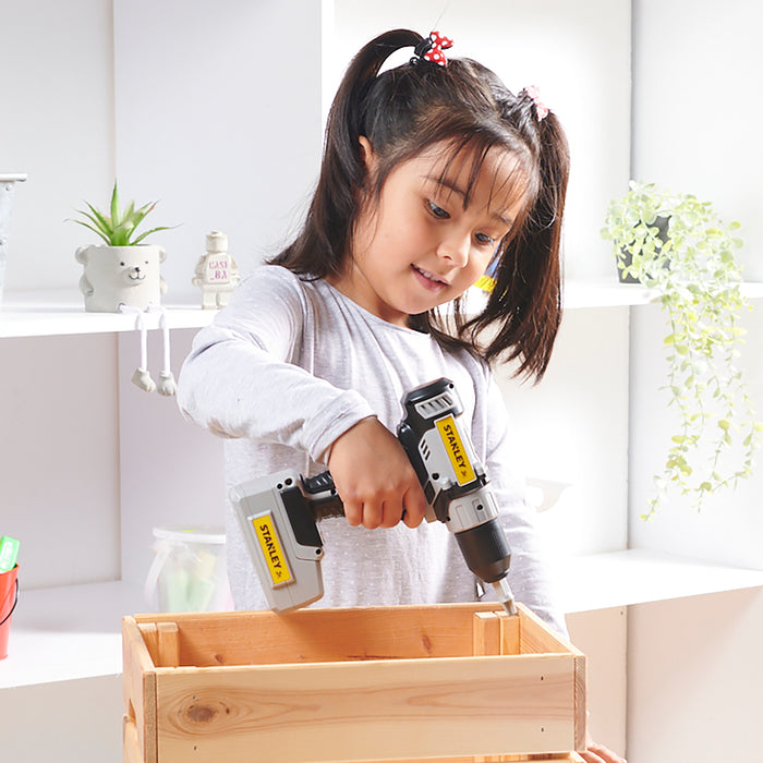 A young girl with pigtails uses a Red Toolbox Stanley Jr. Power Drill to assemble a wooden box in a bright, inviting room, enjoying safe and fun construction play with her kid-friendly power tool.