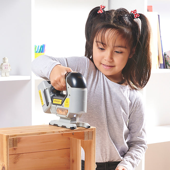 A young girl with pigtails uses the Stanley Jr. Power Tool Set by Red Toolbox to sand a wooden box indoors, smiling and focused, with shelves and books in the backgroundโideal for kidsโ pretend play tool activities.