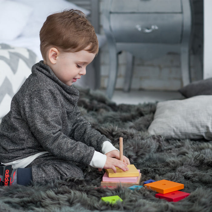 A young child sits on a fluffy gray rug, stacking the colorful Cubika Wooden Pyramid by Cubika. Focused play with this organic wood toy helps develop fine motor skills. A bed and nightstand appear in the background.