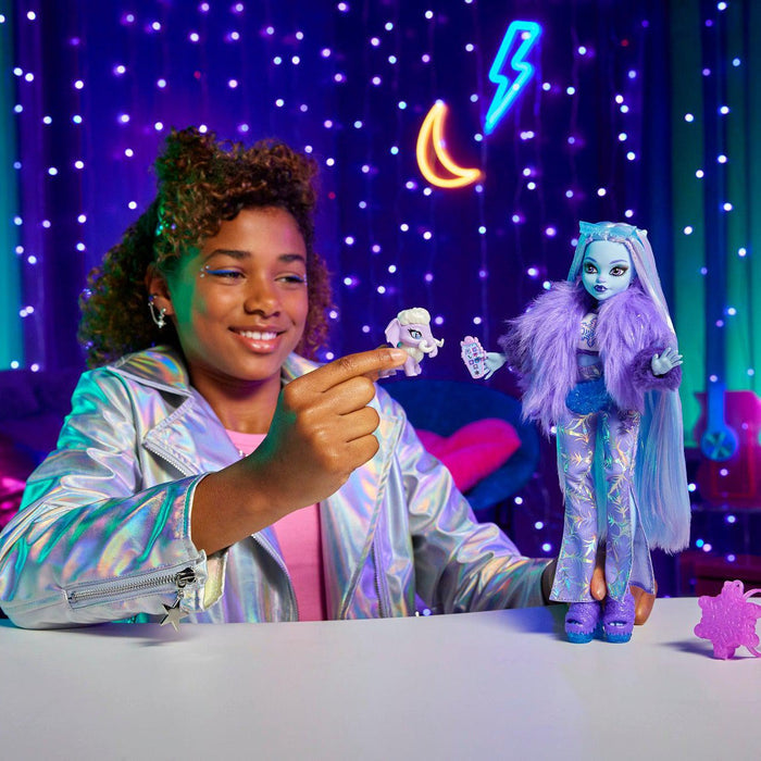 A smiling child in a shiny silver jacket plays with the Mattel Monster High Abbey Doll by Mattel, along with her pet mammoth, seated at a table against a colorful, neon-lit, starry backdrop.
