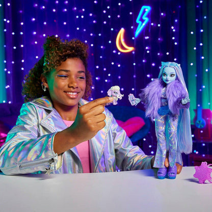A smiling child in a shiny silver jacket plays with the Mattel Monster High Abbey Doll by Mattel, along with her pet mammoth, seated at a table against a colorful, neon-lit, starry backdrop.