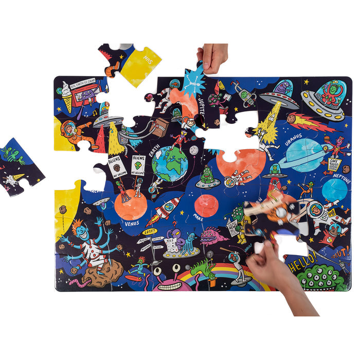 Two hands assemble the Nolja Space 36pc Jigsaw Puzzle by Nolja, depicting planets, spaceships, astronauts, and cartoon characters on a starry background, with several pieces still waiting to be placed.