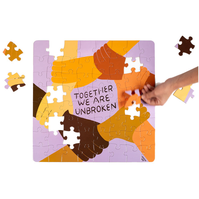 A hand adds a piece to the Nolja Unbroken 64pc Jigsaw Puzzle by Nolja, featuring interlocking hands of diverse skin tones and "TOGETHER WE ARE UNBROKEN" in the center for inspiring, hands-on play.