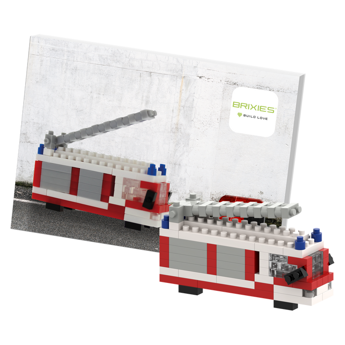 The Brixies Postcard Fire Truck is a LEGO-style mini build kit with a gray ladder, displayed in front of its instruction booklet—a fun, mailable postcard for builders and collectors.