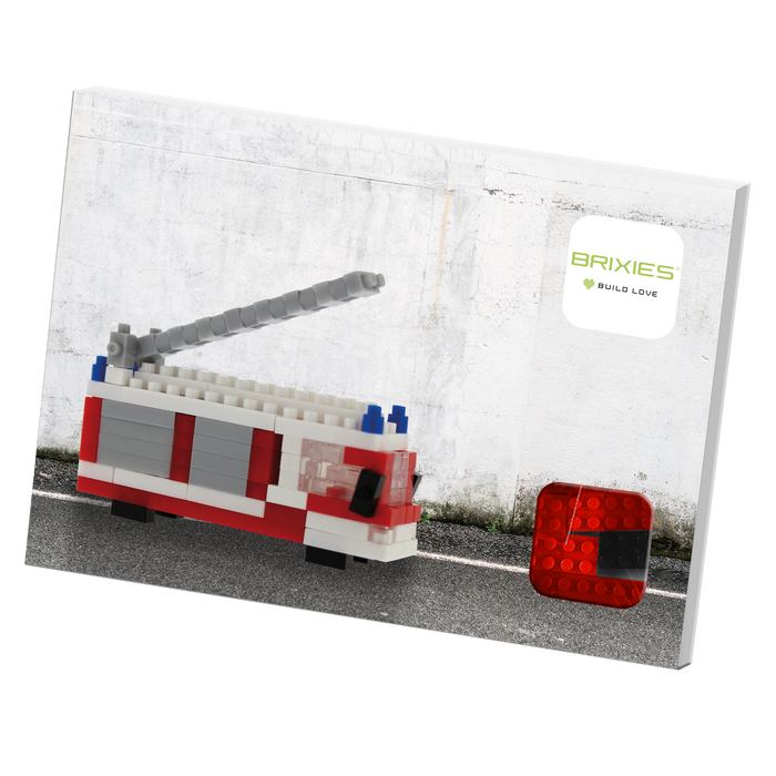A box featuring a BRIXIES Postcard Fire Truck building set made of bricks, displayed against a white wall with the Brixies logo and "Build Love" tagline—perfect as a mailable postcard.