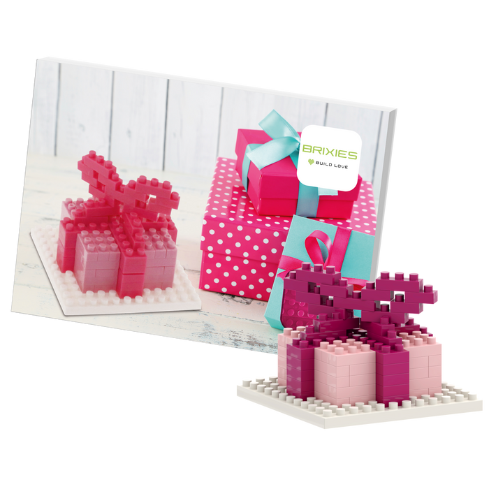 The Brixies Postcard Gift features a small pink and red 3D gift box built from micro bricks, displayed in front of a postcard showing several wrapped presents, including a pink polka-dotted box with a blue ribbon.