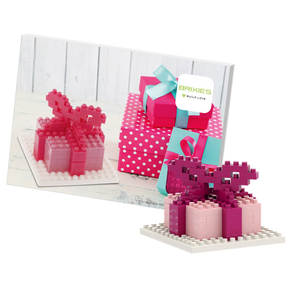 The Brixies Postcard Gift features a small pink and red 3D gift box built from micro bricks, displayed in front of a postcard showing several wrapped presents, including a pink polka-dotted box with a blue ribbon.