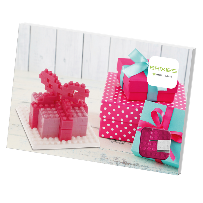 A group of pink and polka dot gift boxes, some with teal ribbons, sits beside a pink BRIXIES Postcard Gift micro-brick model on a white wooden surface. A sign reads "BRIXIES BUILD LOVE," evoking a charming Brixies Postcard Gift scene.