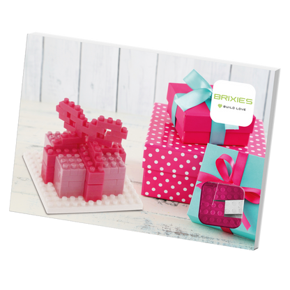 A group of pink and polka dot gift boxes, some with teal ribbons, sits beside a pink BRIXIES Postcard Gift micro-brick model on a white wooden surface. A sign reads "BRIXIES BUILD LOVE," evoking a charming Brixies Postcard Gift scene.