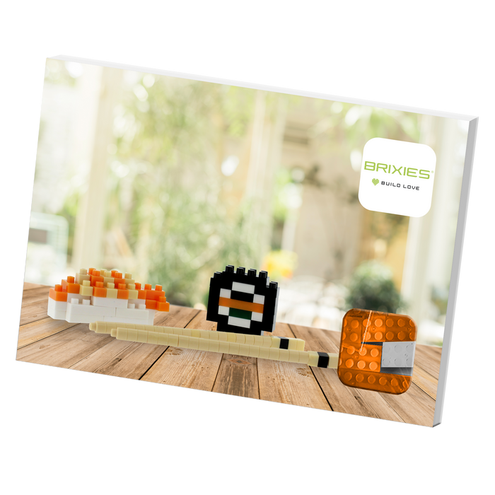 The Brixies Postcard Sushi set features three sushi-themed 3D models made from plastic building blocks, displayed on a wooden surface with a blurred indoor background and the BRIXIES Build Love logo in the top right corner.