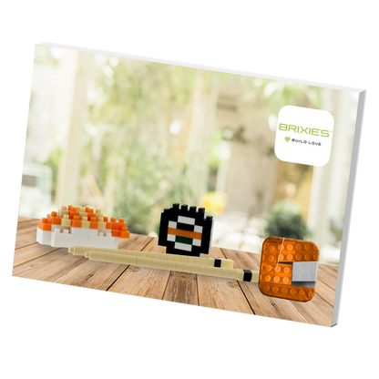The Brixies Postcard Sushi set features three sushi-themed 3D models made from plastic building blocks, displayed on a wooden surface with a blurred indoor background and the BRIXIES Build Love logo in the top right corner.