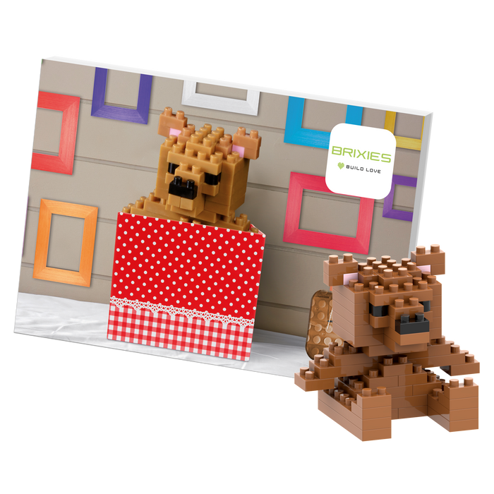 A 3D teddy bear made from interlocking bricks by Brixies, called "Postcard Teddy Bear," sits in a red polka-dot box next to its photo, with colorful frames and a Brixies sign in the background.