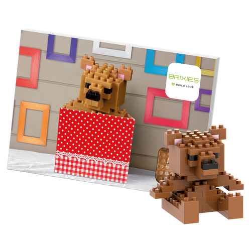A 3D teddy bear made from interlocking bricks by Brixies, called "Postcard Teddy Bear," sits in a red polka-dot box next to its photo, with colorful frames and a Brixies sign in the background.