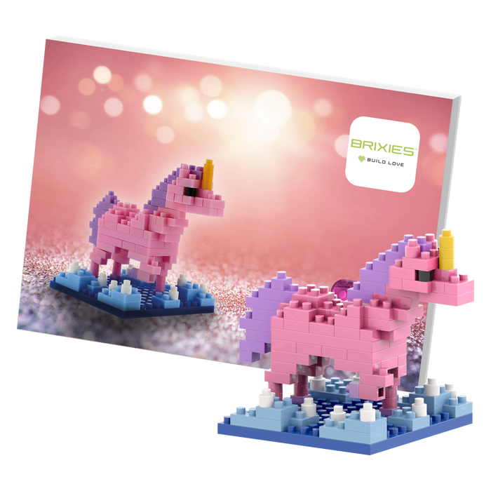 The Brixies Postcard Unicorn is a small pink and purple micro brick figure on a blue base, displayed with matching artwork and the Brixies logo set against a soft pink bokeh background.