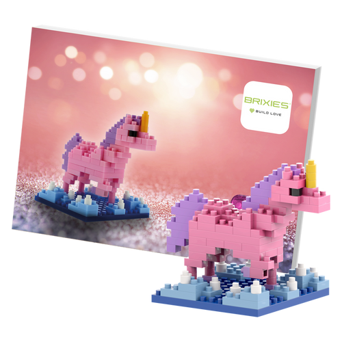 The Brixies Postcard Unicorn is a small pink and purple micro brick figure on a blue base, displayed with matching artwork and the Brixies logo set against a soft pink bokeh background.