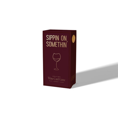 A burgundy box from Som Ventures, labeled “Sippin on Somthin’” with a minimalist wine glass design on the front. “Wine Card Game” is written below, inviting you to play, connect, and learn fun wine trivia. The box casts a rightward shadow.
