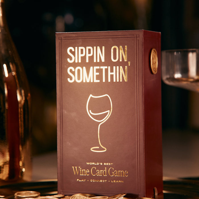 A maroon box from Som Ventures features gold "Sippin on Somthin'" lettering above a wine glass. Below, it states "World's Best Wine Card Game—Play, Connect, Learn," with gold coins and a wine glass for this fun trivia and tasting experience.