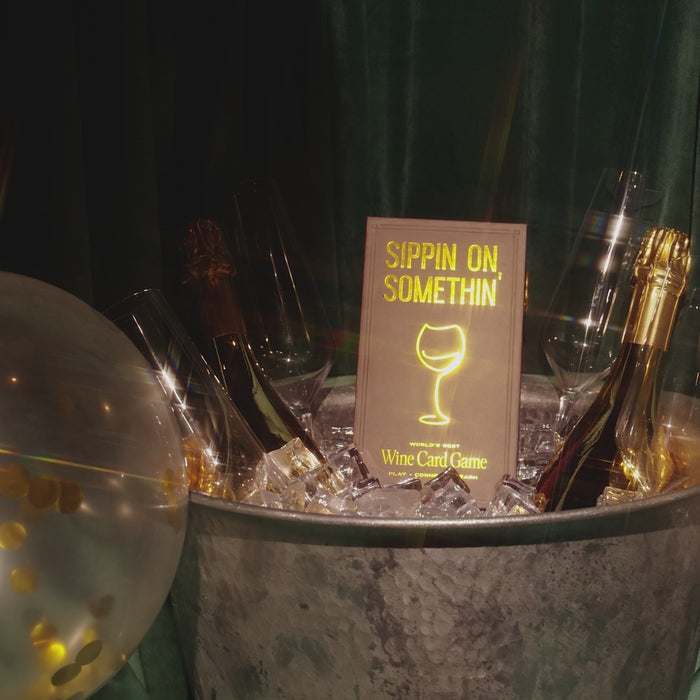 A metal tub with ice, champagne, and glasses sits before a green curtain. A sign reading "Sippin on Somthin'" by Som Ventures with a wine glass illustration—ideal for wine tasting or playing a fun wine trivia game.