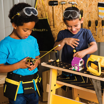 Two young boys in a workshop use the Red Toolbox Stanley Jr. 10 Piece Toolset at a workbench—one measures with a tape measure, the other uses a screwdriver on a model house. Both wear safety goggles and tool belts from the set.