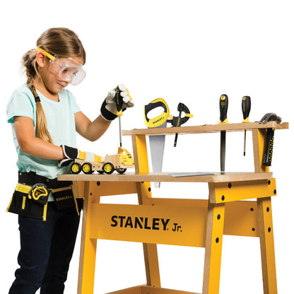 A young girl wearing safety goggles and gloves uses a screwdriver from the Red Toolbox Stanley Jr. 10 Piece Toolset on a toy truck at a yellow workbench with various play tools neatly arranged on top.