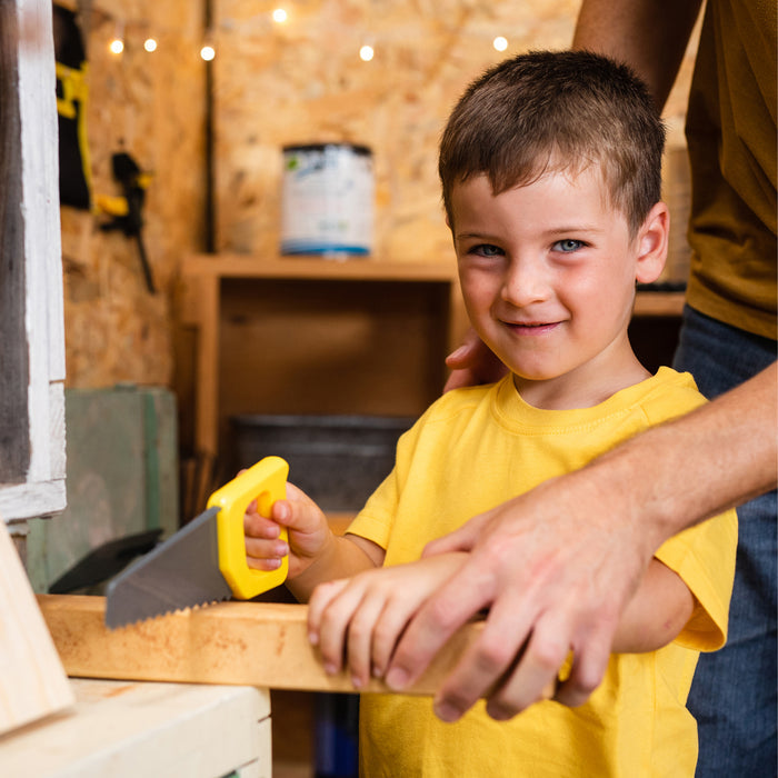 A young boy in a yellow shirt smiles as he uses the Red Toolbox Stanley Jr. 19 Piece Pretend Play Tool Set to cut wood, with an adult's hands assisting him in a cozy workshop setting.