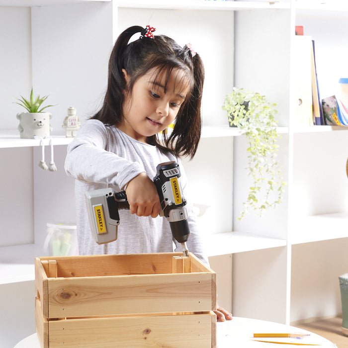 A young girl with pigtails uses the Red Toolbox Stanley Jr. Battery Operated 21 Piece Pretend Play Toolset’s drill to assemble a wooden crate in a bright room with white shelves and plants in the background.