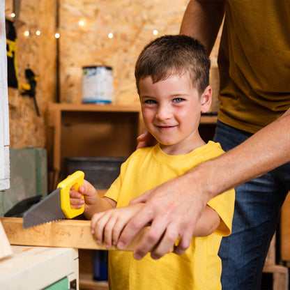 A young boy in a yellow shirt smiles at the camera as he uses the Red Toolbox Stanley Jr. Battery Operated 21 Piece Pretend Play Toolset to cut wood, guided by an adult’s hands in a workshop.