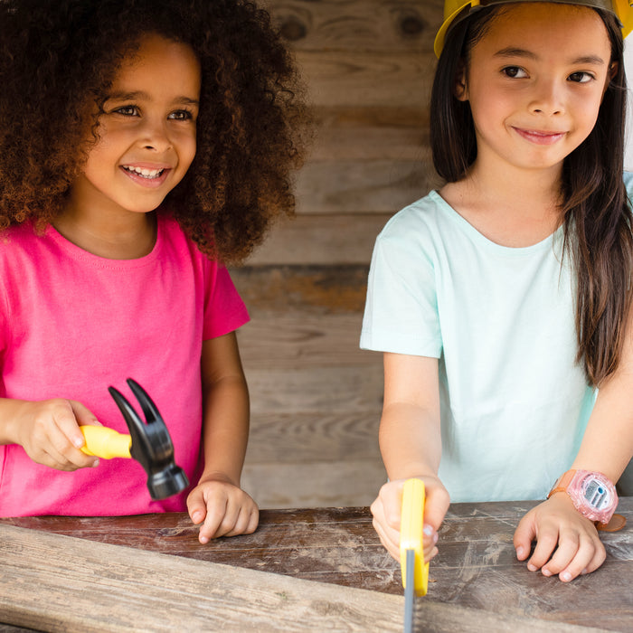 Two young girls smile while using the Red Toolbox Stanley Jr. Battery Operated 21 Piece Pretend Play Toolset; one holds a hammer, the other a screwdriver, both engaged and excited by their creative building activity at a wooden surface.