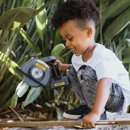 A young child in a white shirt and jeans smiles while playing with the Stanley Jr. Power Tool Set by Red Toolbox on a wooden plank outdoors, surrounded by lush green plants—perfect for kids’ pretend play tool fun.