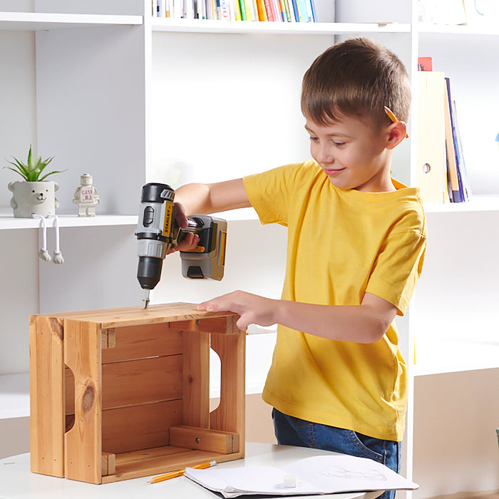 A young boy in a yellow t-shirt smiles as he uses the Red Toolbox Stanley Jr. Power Tool Set to assemble a wooden crate, enjoying the fun of battery-operated toy tools.
