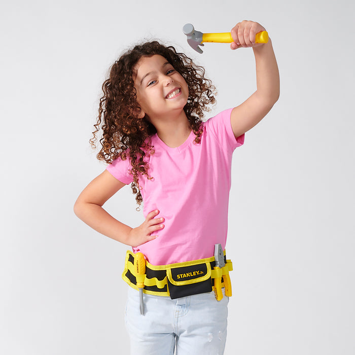 A smiling girl with curly hair in a pink shirt and the Stanley Jr. Pretend Play Tools & Toolbelt Set by Red Toolbox playfully holds a yellow toy hammer above her head, posing confidently against a plain background.