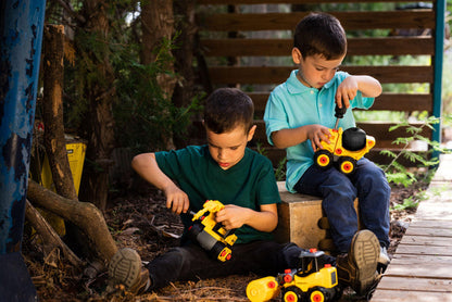 Two young boys play outdoors with the Red Toolbox Stanley Jr. Take Apart Classic Cement Mixer and other construction vehicles. One in a green shirt sits on the ground; another in blue perches on a wooden box, with trees and a fence behind them.