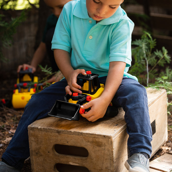 A boy in a blue shirt plays outdoors with the Red Toolbox Stanley Jr. Take Apart Classic Front Loader, sitting on a wooden box—the ideal toy for young aspiring builders.
