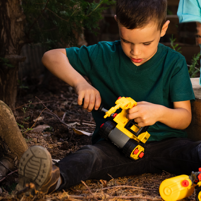 A young boy in a green shirt sits outdoors, immersed in play with the Red Toolbox Stanley Jr. Take Apart Classic Mega Construction Bundle, using a screwdriver to assemble parts and enjoying his STEM kit experience amid dirt and greenery.