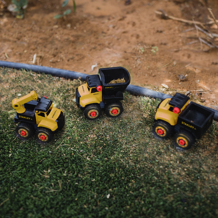 Three small yellow vehicles—a dump truck, crane, and another dump truck—from the Stanley Jr. Take Apart Dot 5 Piece Set with Battery Operated Screwdriver by Red Toolbox are placed on grass by a dirt patch, perfect for hands-on play for little builders.
