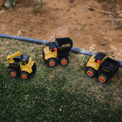 Three small yellow vehicles—a dump truck, crane, and another dump truck—from the Stanley Jr. Take Apart Dot 5 Piece Set with Battery Operated Screwdriver by Red Toolbox are placed on grass by a dirt patch, perfect for hands-on play for little builders.