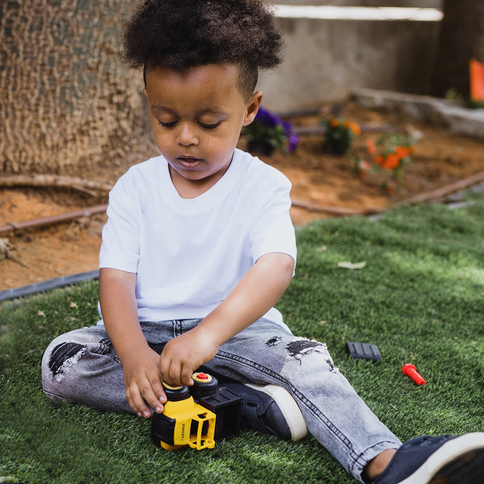 A young child with curly hair, dressed in a white t-shirt and ripped jeans, plays on artificial grass with the Red Toolbox Stanley Jr. Take Apart Dot 5 Piece Set & Battery Operated Screwdriver, surrounded by toy parts, a tree, and flowers.