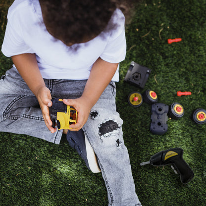 A child sits on grass wearing a white shirt and ripped gray jeans, playing with small toy vehicles and tool pieces from the Red Toolbox Stanley Jr. Take Apart Dot 5 Piece Set & Battery Operated Screwdriver STEM kit nearby.