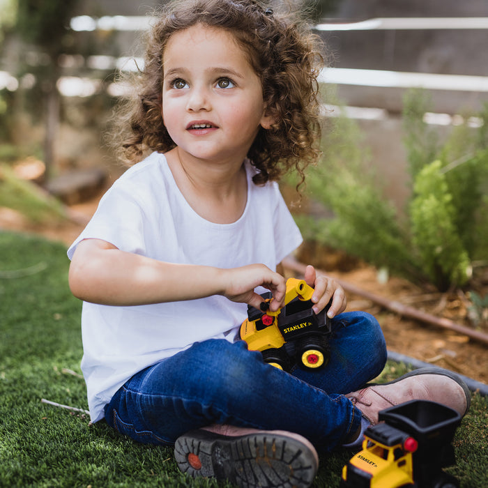 A young child with curly hair, wearing a white shirt and blue jeans, sits on the grass outdoors playing with the Red Toolbox Stanley Jr. Take Apart Dot 5 Piece Set & Battery Operated Screwdriver, looking up thoughtfully.