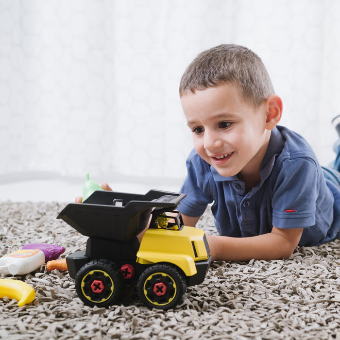 A young boy smiles while playing on a carpeted floor with the Stanley Jr. Take Apart XL Dump Truck by Red Toolbox and colorful plastic fruits and vegetables, as a white patterned curtain hangs in the background.