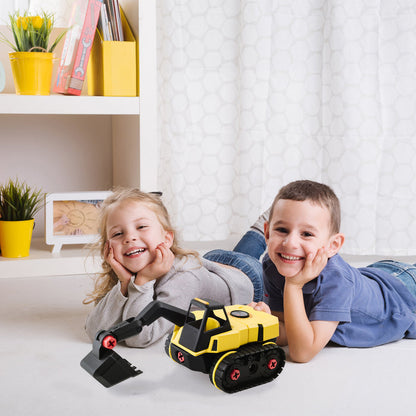 Two smiling children lie on the floor indoors, propped up beside a yellow Stanley Jr. Take Apart XL Excavator construction toy by Red Toolbox. A white shelf with plants, books, and framed photos is in the background.