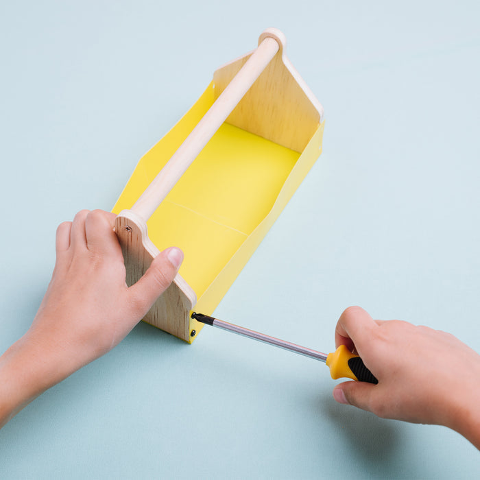 Using a screwdriver, a person assembles the Stanley Jr. Toolbox Kit by Red Toolbox, attaching a yellow side panel to the base of the wooden DIY kit.