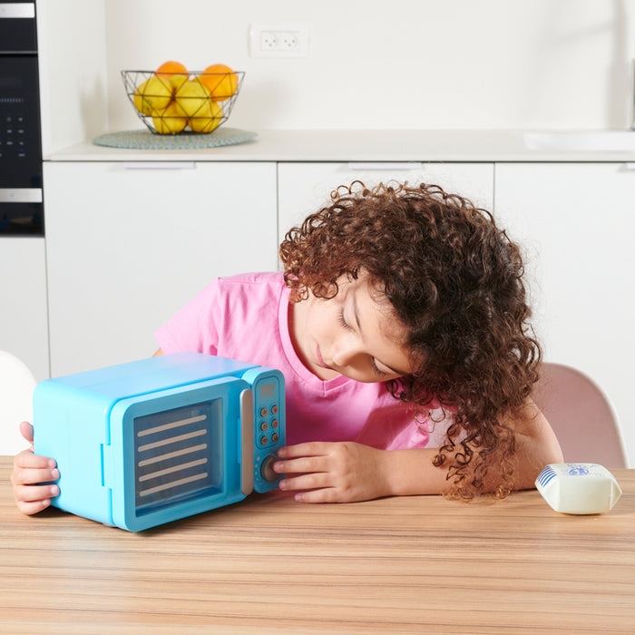 A child in a pink shirt plays with the Red Toolbox Tasty Junior Pretend Play Microwave Set, pressing its blue buttons and surrounded by pretend food. A bowl of fruit sits on a counter in the background.
