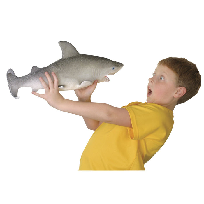 A surprised boy in a yellow shirt holds the Toysmith Ginormous Grow Shark water-grow toy with both hands, staring at it wide-eyed and open-mouthed against a plain white background.